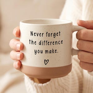 Può includere: Una tazza in ceramica color crema con finitura maculata e una base solida color terracotta. La parte anteriore presenta il testo stampato in nero 'Never forget the difference you make.' seguito da un piccolo cuore disegnato a mano.