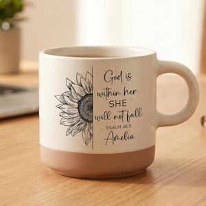 May include: Cream ceramic mug with a brown base, decorated with a black and white sunflower and the text "God is within her. SHE will not fall" and the name "Amelia". The mug is on a wooden surface.