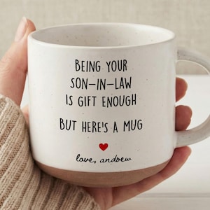 Könnte beinhalten: Weißer Keramikbecher mit braunem Boden, mit dem schwarzen Text "BEING YOUR SON-IN-LAW IS GIFT ENOUGH BUT HERE'S A MUG". Ein kleines rotes Herz befindet sich unter dem Text, darunter steht "love, andbew".