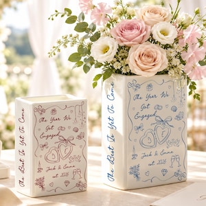 May include: Two white rectangular ceramic vases featuring line art illustrations of two hearts with initials J and E, the text The Year We Got Engaged, Jack & Emma, est. 2026, and The Best Is Yet To Come. One vase has red ink, the other blue ink.