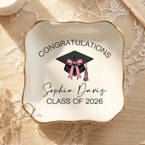 May include: A cream-coloured ceramic trinket dish with a gold edge, inscribed with "CONGRATULATIONS" and "Sophia Davis CLASS OF 2026". A graduation cap with a pink bow is in the centre. A ring rests on the dish.