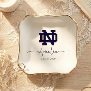 May include: A white ceramic trinket dish with a gold rim, featuring a navy blue logo and the name "Amelia" with "Class of 2026" below. A ring rests on the edge. The dish is on a lace-covered surface.