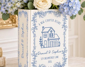 May include: A white ceramic book-shaped vase with blue floral borders and a house illustration. Text reads: A NEW CHAPTER BEGINS, Daniel & Sophia, 524 DREAMSTREET LANE, 2024. It holds a bouquet of white roses, blue hydrangeas, and delphiniums.