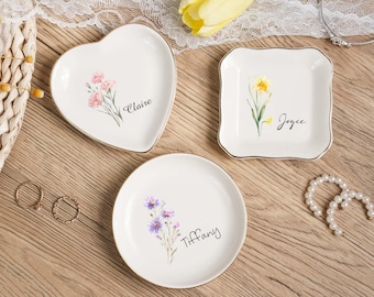 May include: Three white ceramic trinket dishes with gold rims. One is heart-shaped, one square, and one round. Each features a floral design and a name: Claire, Joyce, and Tiffany. The dishes are on a wooden surface.