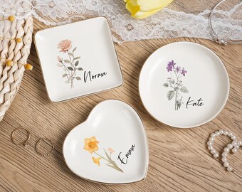 May include: Three white ceramic trinket dishes with gold rims. One square, one round, and one heart-shaped. Each features a floral design and a name: Norma, Kate, and Emma. Rings and a pearl necklace are also visible.
