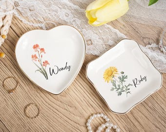 May include: Two white ceramic trinket dishes with gold rims. One is heart-shaped with orange floral accents and the name "Wendy". The other is square with a yellow flower and the name "Ruby".