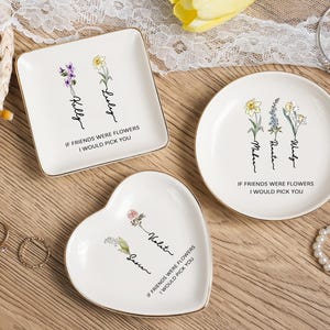 Può includere: Tre piatti portagioie in ceramica bianca con bordi dorati. Uno quadrato, uno rotondo e uno a forma di cuore. Ognuno presenta illustrazioni floreali e il testo "If friends were flowers I would pick you" con nomi. Sono visibili anche anelli e una collana di perle.
