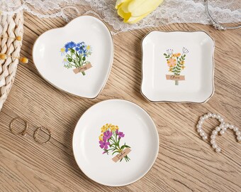 May include: Three white ceramic trinket dishes with gold rims. One is heart-shaped, one square, and one round. Each features a floral bouquet design with a name label. The heart-shaped dish says "Daisy". The square dish says "Olivia". The round dish says "Sophia".