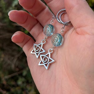 May include: Silver earrings with a star and spiral design. The earrings feature a clear, blue and silver bead and a dangling star-shaped charm. The earrings are held in a hand.