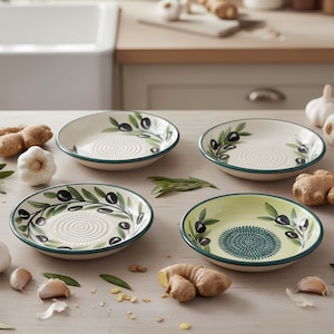 Może przedstawiać: Cztery ceramiczne miseczki na oliwę z powierzchnią do ścierania. Miseczki mają białą podstawę, zielone obramowanie i wzory gałązek oliwnych. Czosnek i imbir są rozrzucone wokół misek na jasnej drewnianej powierzchni.