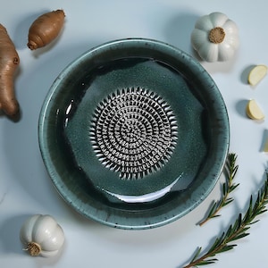 Puede incluir: Un cuenco rallador de cerámica turquesa con una superficie de rallado texturizada en el centro. El cuenco está rodeado de ingredientes frescos: jengibre, batata, dientes de ajo, ramitas de romero y un bulbo de ajo entero.