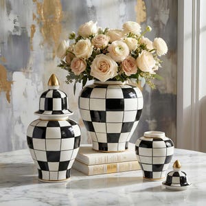 Könnte beinhalten: Eine Sammlung dekorativer Keramikvasen und -gläser mit einem schwarz-weißen Schachbrettmuster. Eine Vase enthält einen Strauß hellrosa und cremefarbener Rosen. Die Gläser haben goldene Akzente.
