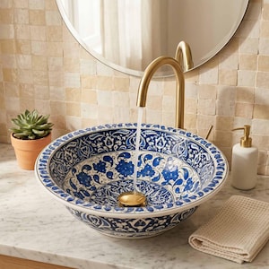 Puede incluir: Un lavabo de cerámica redondo con un patrón floral azul y blanco, combinado con un grifo alto de oro cepillado. El lavabo está sobre una encimera de mármol blanco junto a una pequeña planta suculenta y un dispensador de jabón blanco.