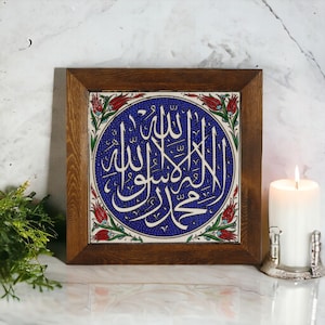 May include: Square wooden framed tile with blue and white calligraphy. The calligraphy is centered on the tile, surrounded by a white border with red and green floral accents. A lit white candle sits to the right.