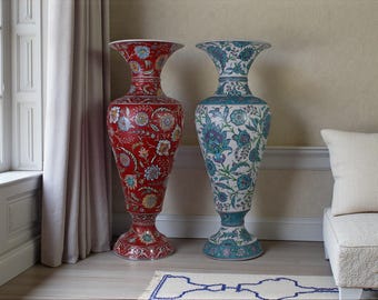 May include: Two tall decorative vases. One is red with floral patterns, the other is white with blue and green floral designs. Both have flared rims and ornate bases. They stand on a light wood floor.
