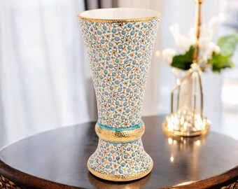 Può includere: Un vaso decorativo in ceramica con un motivo floreale ripetuto in turchese e giallo su sfondo bianco. Il vaso presenta finiture dorate. È posto su un tavolo di legno scuro. Il vaso misura circa 25 cm di altezza.