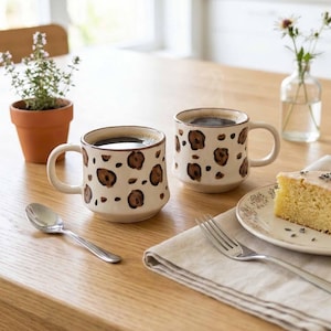 May include: Two ceramic coffee mugs filled with dark liquid, featuring a cream-colored base and brown leopard print spots. A spoon, fork, and a slice of cake are on a wooden table, alongside a small potted plant and a vase of flowers.