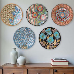 Peut inclure: Cinq assiettes décoratives en céramique avec des motifs floraux colorés sont accrochées au mur. Les assiettes présentent divers motifs, notamment des fleurs, des arbres et des formes géométriques. Un meuble en bois avec des vases et des livres se trouve en dessous.