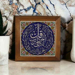 May include: Square wooden framed tile with a blue circular design featuring white Arabic calligraphy. The tile is surrounded by a decorative border with red, yellow, and green floral patterns. The frame is a warm brown color.