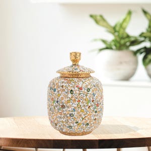 Puede incluir: Un tarro decorativo de cerámica con tapa, con un fondo blanco adornado con coloridos diseños florales. El tarro tiene ribetes dorados y un remate dorado en la tapa. El tarro está sobre una mesa de madera.