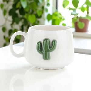 May include: White ceramic mug with a green cactus design. The mug has a rounded shape with a handle and a raised cactus applique on the front. The mug is sitting on a white surface.
