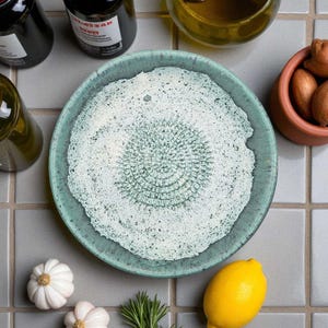 Puede incluir: Un plato rallador de cerámica turquesa con una superficie texturizada, rodeado de ingredientes como ajo, un limón y romero. El plato tiene una zona de rallado central y circular. También se ven varias botellas y un cuenco pequeño de terracota.