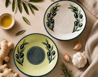May include: Two round ceramic grater plates featuring hand-painted olive branch motifs. One plate is cream-colored with a white center grater, while the other is light green with a dark green center grater. Both have dark green rims.