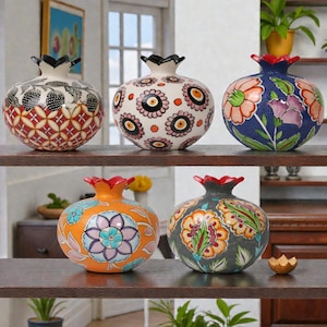 Puede incluir: Cinco jarrones decorativos de cerámica con forma de granada y complejos diseños florales y geométricos. Los jarrones se exhiben en estantes de madera oscura, mostrando una variedad de colores, incluyendo rojo, naranja, azul y blanco. Cada jarrón tiene un diseño único.
