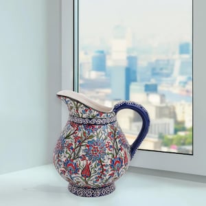 Pode incluir: Jarro de cerâmica com uma alça azul e um bico branco. O jarro é decorado com um padrão floral em vermelho, azul e verde sobre um fundo branco. Está colocado sobre uma superfície branca perto de uma janela.
