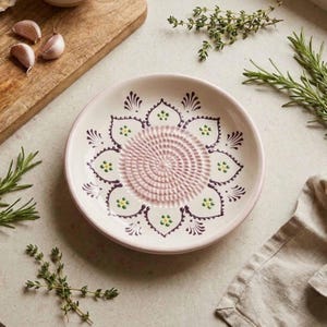 Puede incluir: Un plato rallador de ajo de cerámica con diseño floral. El plato es blanquecino con centro y borde rosa, y presenta un patrón decorativo en morado y verde. Hierbas frescas y dientes de ajo están dispuestos alrededor del plato.