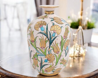 Può includere: Un vaso decorativo bianco con un motivo floreale con dettagli oro, giallo, blu e verde. Il vaso ha una forma arrotondata e un collo stretto, con finiture dorate. È posizionato su un tavolo marrone scuro. Misura circa 30 cm di altezza.