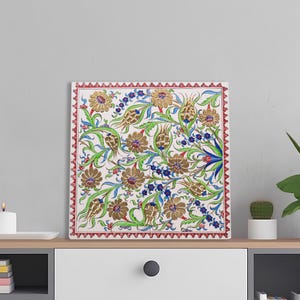 May include: Square floral art print featuring a detailed pattern of stylized flowers and foliage in gold, red, blue, and green on a cream background. The print has a red border with a repeating triangular design. The artwork is displayed on a white surface.