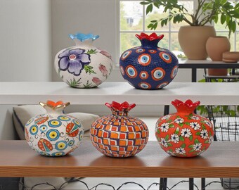 May include: Decorative ceramic pomegranate-shaped vases. The vases feature intricate designs, including floral patterns, geometric shapes, and eye motifs. The color palette includes white, blue, orange, and red. These vases are ideal for home decor.