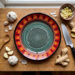 Puede incluir: Un plato rallador de ajo de cerámica con un diseño rojo y verde, rodeado de dientes de ajo, una cuchara de madera y un cuenco de jengibre rallado. También hay un cuchillo y raíz de jengibre, lo que sugiere uso culinario.