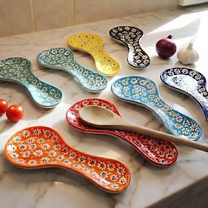 May include: A collection of colorful ceramic spoon rests with a floral daisy pattern. The rests are in various colors, including orange, red, yellow, blue, and teal. A wooden spoon rests on a red spoon rest. Vegetables are also present.