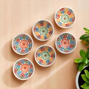 May include: Six small, white ceramic bowls with colorful floral patterns in red, orange, blue, and green. The bowls are arranged on a light wooden surface, with a plant in the background. Each bowl has a diameter of approximately 4 inches.