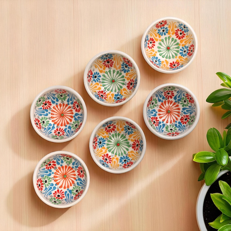 May include: Six small, white ceramic bowls with colorful floral patterns in red, orange, blue, and green. The bowls are arranged on a light wooden surface, with a plant in the background. Each bowl has a diameter of approximately 4 inches.