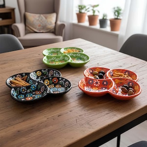 May include: A wooden table displays several colorful, divided serving dishes. The dishes are in green, black, and orange, each with a floral design. Some dishes contain snacks like olives and crackers, while others are empty.