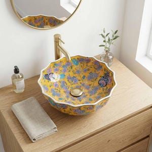 May include: A yellow floral ceramic bathroom sink with a scalloped edge. The sink has a gold tap and drain, and is decorated with blue and pink flowers. A folded beige towel and a bottle of soap sit on the wooden vanity unit.