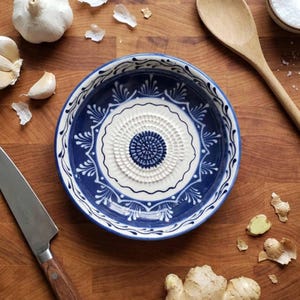 Puede incluir: Un cuenco rallador de ajo de cerámica azul y blanco con un diseño floral decorativo. El cuenco tiene una superficie de rallado texturizada en el centro. Ajos, un cuchillo y una cuchara de madera están sobre una superficie de madera.