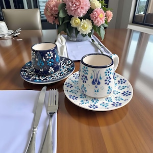 May include: Two ceramic coffee cups and saucers with blue floral patterns. One cup is navy blue, the other is white. The saucers and cups have a matching design. A knife and fork are on a white napkin.