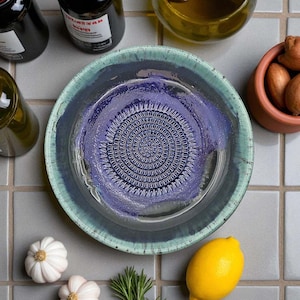 Puede incluir: Un cuenco rallador de cerámica hecho a mano con una superficie de rallado circular texturizada en el centro. El cuenco tiene un borde azul-verdoso y un interior morado. Ajo, un limón y otros ingredientes rodean el cuenco.