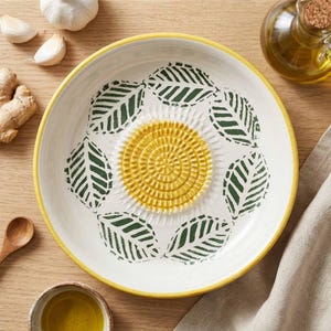 Może przedstawiać: Biała ceramiczna tarka do czosnku z żółtym środkiem i obramowaniem oraz wzorem zielonych liści. Tarka otoczona jest składnikami takimi jak czosnek, imbir i oliwa z oliwek.