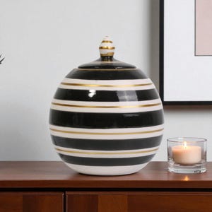 Puede incluir: Un jarrón decorativo de cerámica con tapa, con rayas horizontales negras y blancas con detalles dorados. El jarrón está sobre una superficie de madera, con una vela encendida en un portavelas de cristal.