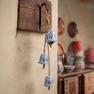 May include: Three ceramic bells with blue floral patterns hang from a wooden plaque. The bells are suspended by a rope with colourful beads. The background shows other decorative items.