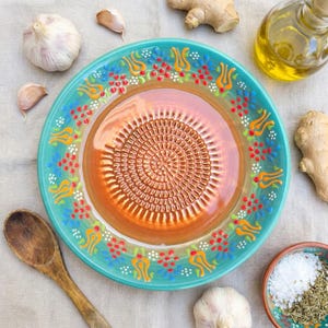 Może przedstawiać: Turkusowy ceramiczny talerz do ścierania z kwiatowym wzorem na obrzeżu. Środek talerza to teksturowana, spiralna powierzchnia do ścierania. Czosnek, korzeń imbiru, oliwa z oliwek i drewniana łyżka otaczają talerz.