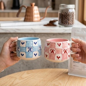 May include: Two ceramic mugs, one light blue and one pink, are held in hands. Each mug features a checkered pattern with bows and hearts. The mugs have white handles and interiors, perfect for coffee or tea.