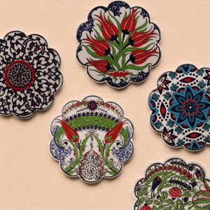 May include: A collection of five decorative ceramic coasters with scalloped edges. Each coaster features a unique floral or geometric design in red, blue, green, and white against a cream background. The coasters are likely for home decor.