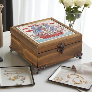 May include: A wooden jewelry box with a decorative tile top featuring a ship design in blue, red, and white. The box has metal accents and feet. Jewelry trays with necklaces and rings are in the foreground.
