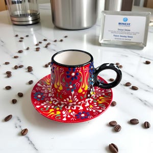 May include: A small, hand-painted ceramic coffee cup and saucer set. The cup and saucer are red with a floral design in blue, yellow, and white. The cup has a black handle with white polka dots. Coffee beans are scattered around the set.